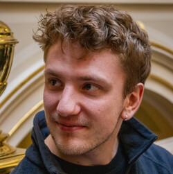 Julian Schmiedel hat kurze, blonde gelockte Haare und einen Drei-Tage-Bart. Er trägt einen blauen Fleece-Pullover und einen silbernen Ohrring. 