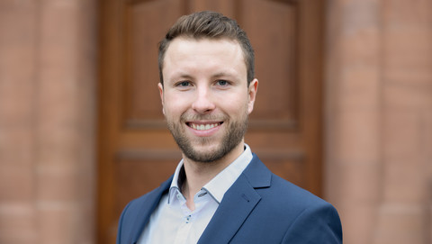 Philip Holler. He is wearing a blue suit and is standing in front of a brown door and wall.