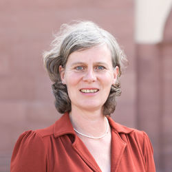Portrait von Ursula Schlichter. Sie trägt eine rote Bluse und steht lächelnd vor dem Schlossgebäude.