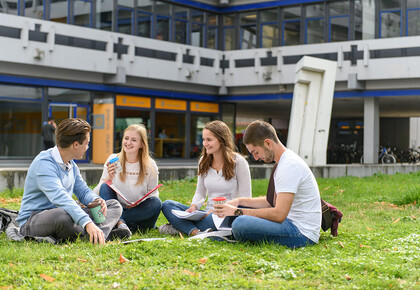 Vier Studierende sitzen mit Ordnern und Kaffee auf einer Wiese vor einem Uni-Gebäude