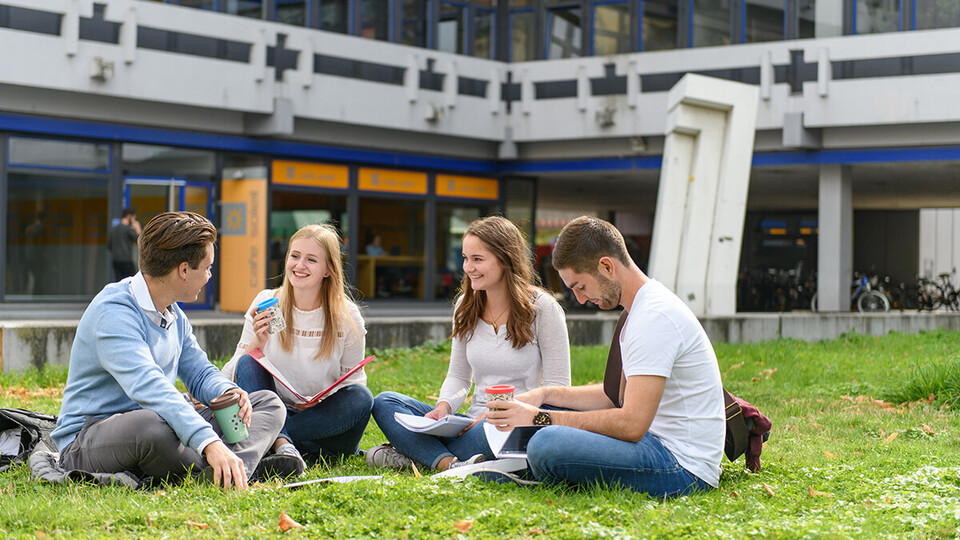 Vier Studierende sitzen mit Ordnern und Kaffee auf einer Wiese vor einem Uni-Gebäude