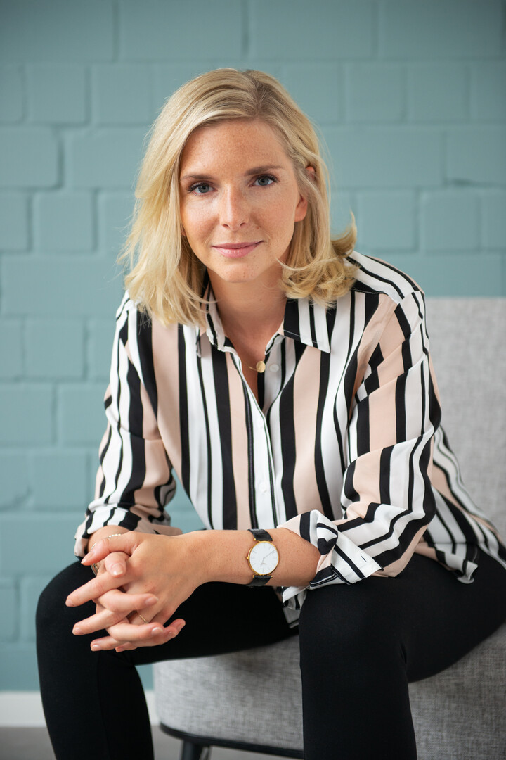  Lea-Sophie Cramer sitzt auf einem Sofa. Sie trägt eine schwarze Hose sowie eine weiß-schwarz-rosa-karriertes Bluse.