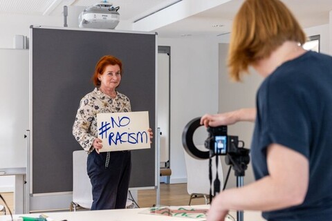 Person macht mit Kamera Foto von einer Frau, die ein Schild mit der Aufschrift "No Racism", in der Hand hält.