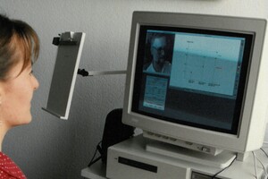 Eine Frau in roter Bluse arbeitet an einem alten Computer, auf dem eine andere Person und Vorlesungsfolien zu sehen sind.