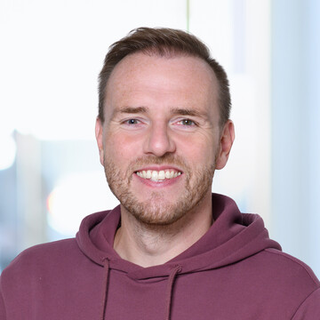 Porträt von Patrick Kullmann. Er trägt einen weinroten Adidas-Pullover und lächelt in die Kamera.
