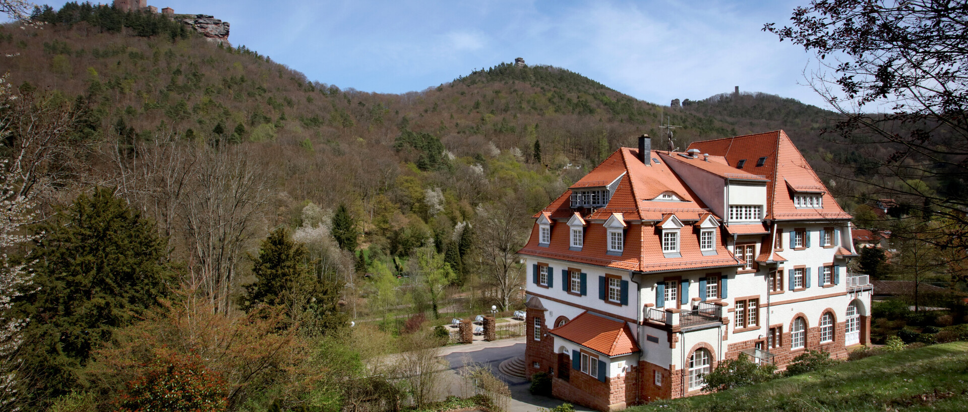 Das Kurhaus Trifels (Tagungshotel) in Annweiler ist im Vordergund zu sehen. Im Hintergrund sieht Bäume aus dem Pfälzer Wald und auf einem Hügel noch angedeutet die Trifelser Burg.