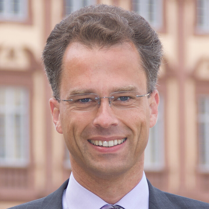 Portrait von Georg Bitter. Er tträgt ein weißes Hemd, eine lila-farbene Krawatte sowie einen grauen Blazer und steht vor dem Schloss.