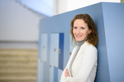 Porträt von Karin Hoisl. Sie hat braune, halblange Haare, trägt ein weißes Jacket und lächelt in die Kamera. Sie lehnt an einer Wand mit blauen Schließfächern.
