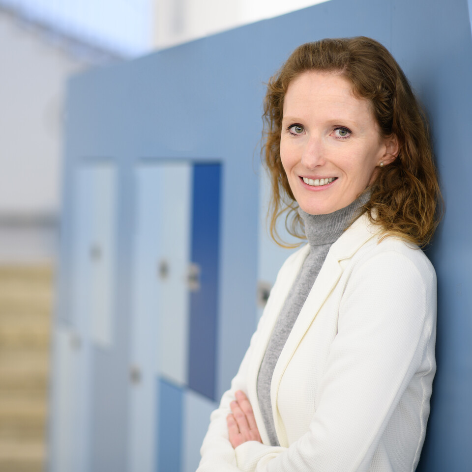 Porträt von Karin Hoisl. Sie hat braune, halblange Haare, trägt ein weißes Jacket und lächelt in die Kamera. Sie lehnt an einer Wand mit blauen Schließfächern.