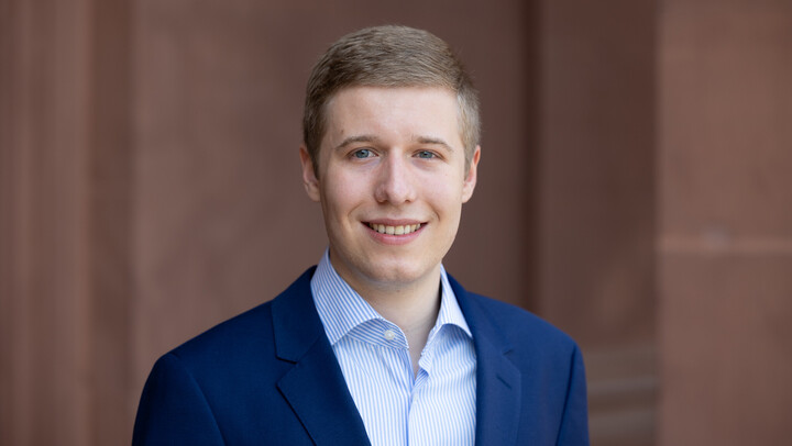 Marc Mertens. He is wearing a blue suit and is standing in front of a brown stone wall.