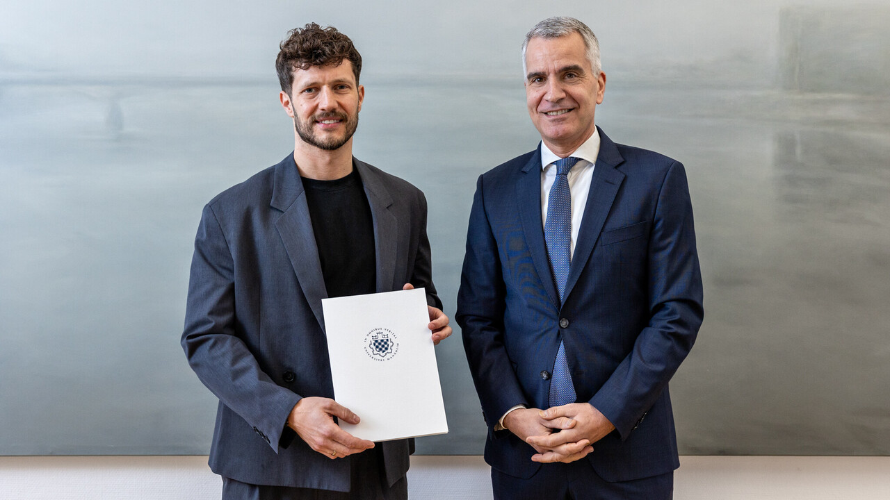 Prof. Olbert (li.) mit der Ernennungsurkunde und Uni-Rektor Prof. Fetzer. Prof. Olbert und Prof. Fetzer stehen nebeneinander. Sie tragen beide dunkle Anzüge und lächeln in die Kamera. Olbert hält die Ernennungsurkunde in der Hand.