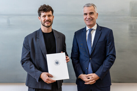 Prof. Olbert und Prof. Fetzer stehen nebeneinander. Sie tragen beide dunkle Anzüge und lächeln in die Kamera. Olbert hält die Ernennungsurkunde in der Hand.