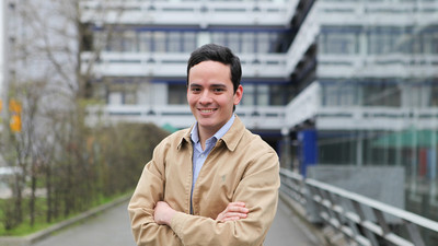 Eine lächelnde Person trägt ein blau-weiß gestreiftes Hemd sowie eine hellblaue Jacke und steht vor dem A5-Gebäude. Die Person heißt Sebastián Echeverría Botero.