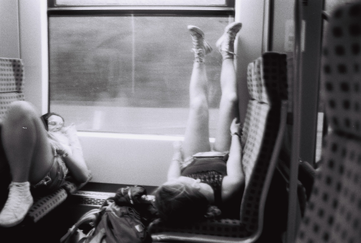 Zwei Frauen liegen auf Sitzen in einem Zug, eine der Frauen hat ihre Beine gegen die Schreibe gedrückt. Das Foto ist in Schwarz Weiß.