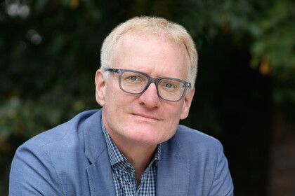 Portrait von Matthias Kohring. Er trägt einen blauen Blazer und sitzt im Freien auf einer Bank.