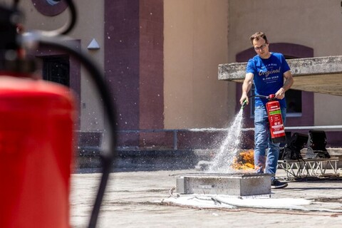 Mann löscht im Hintergrund mithilfe eines Feuerlöschers ein kleines Feuer im Schneckenhof. Im Vordergrund ein Feuerlöscher.