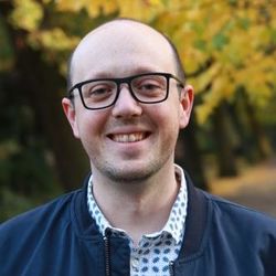 Ein Porträt von Tristan Klingelhöfer. Er trägt eine auffällige schwarze Brille, ein grün-weiß gemustertes Hemd und darüber eine dunkelblaue Jacke.