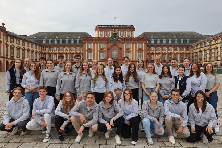 Eine große Gruppe Studierender steht im Ehrenhof. Alle tragen graue Pullis oder Shirts mit dem Fachschaftslogo darauf und lächeln in die Kamera.