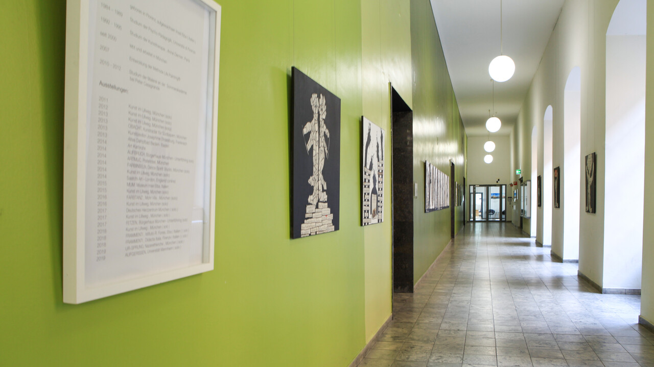 Ein Flur, der mit Kunstwerken beschmückt ist. Links im Vordegrund hängt ein Informationsplakat einer Künstlerin.