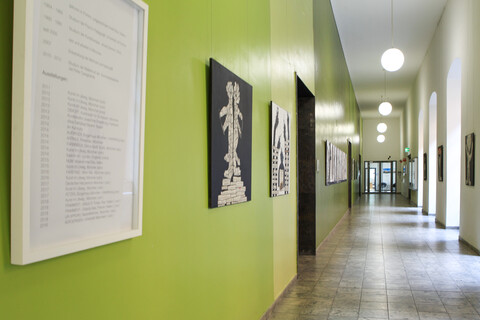 Ein Flur, der mit Kunstwerken beschmückt ist. Links im Vordegrund hängt ein Informationsplakat einer Künstlerin.