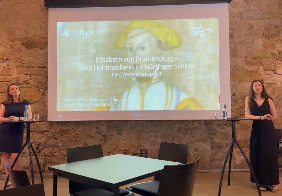 Zwei Frauen stehen an zwei Stehtischen neben einer Leinwand mit der Präsentation 'Elisabeth von Brandenburg – eine Hohenzollern im Nürtinger Schloss' in einem gemauerten Kellergewölbe