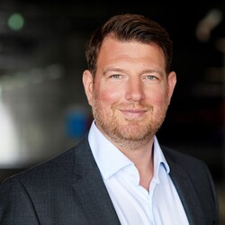 Philipp Laux hat braune Haare und einen grau-braunen Dreitagebart. Er trägt ein graues Jacket und ein weißes Hemd.