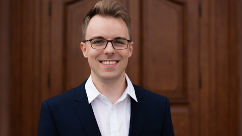 Bennet Janssen. He is wearing a black suit and is standing in front of one of the castle doors.