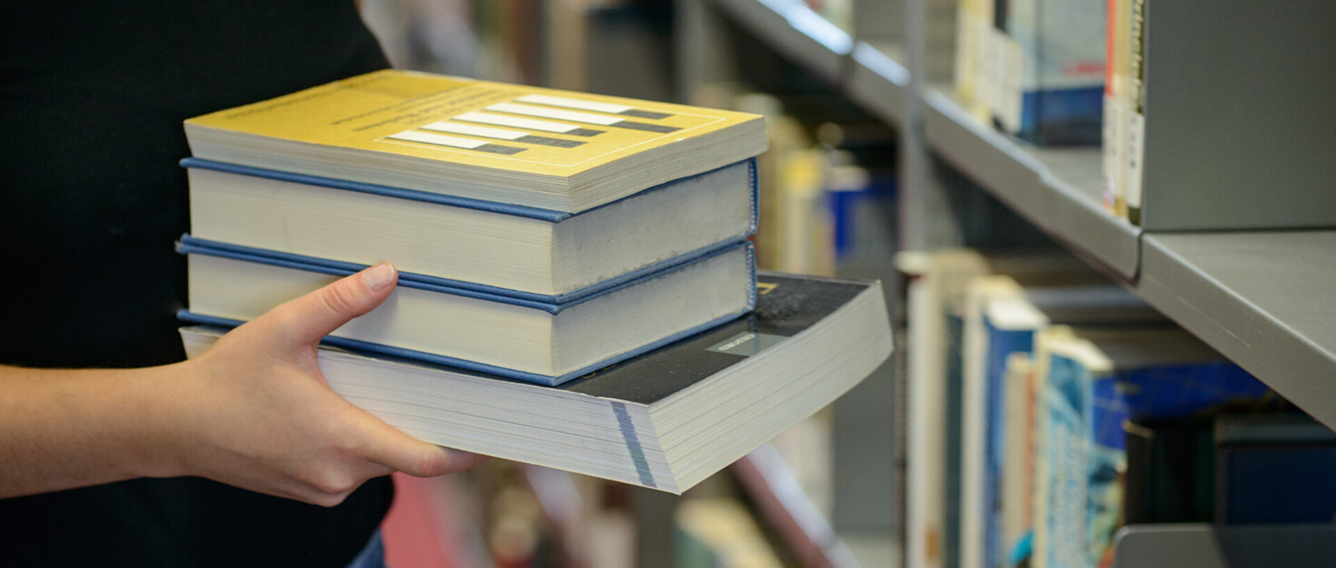 Eine Person hält Bücher in der Hand und im Hintergrund ist ein Bücherregal der Universitätsbibliothek zu sehen.