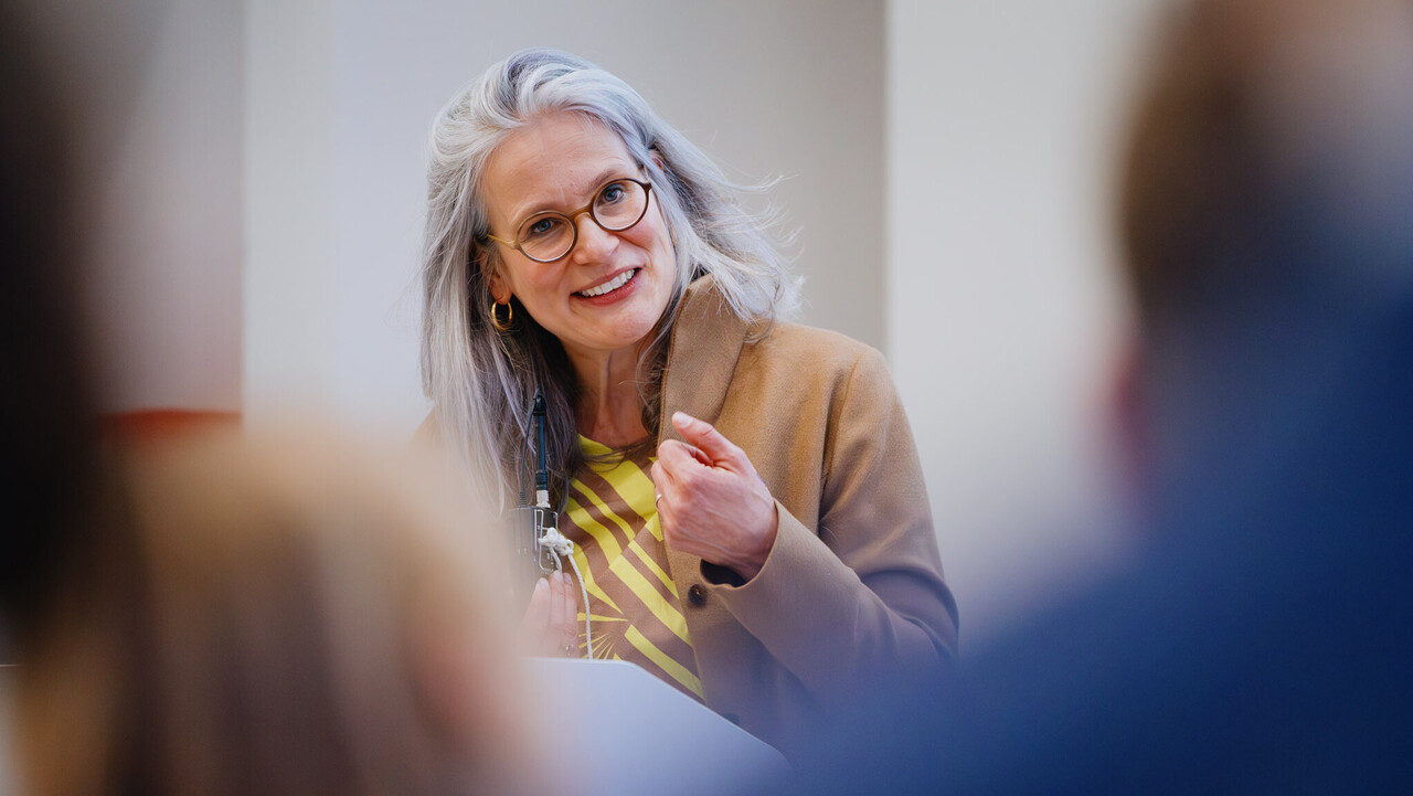 Das Bild zeigt die Prorektorin für Forschung der Universität. Sie steht am Rednerpult und blickt in die Kamera. Im Vordergrund zwei verschommene Köpfe aus dem Publikum. Sie hat graue lange Haaare und ein kamelfarbenes Kostüm an.