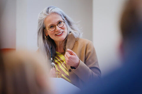 Das Bild zeigt die Prorektorin für Forschung der Universität. Sie steht am Rednerpult und blickt in die Kamera. Im Vordergrund zwei verschommene Köpfe aus dem Publikum. Sie hat graue lange Haaare und ein kamelfarbenes Kostüm an.