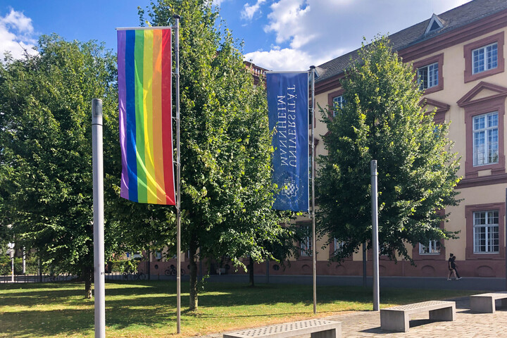 Die LSBT*QIA-Flagge in bunten Regenbogenfarben und die blaue Flagge mit der Aufschrift "Universität Mannheim" vor dem Schloss. 