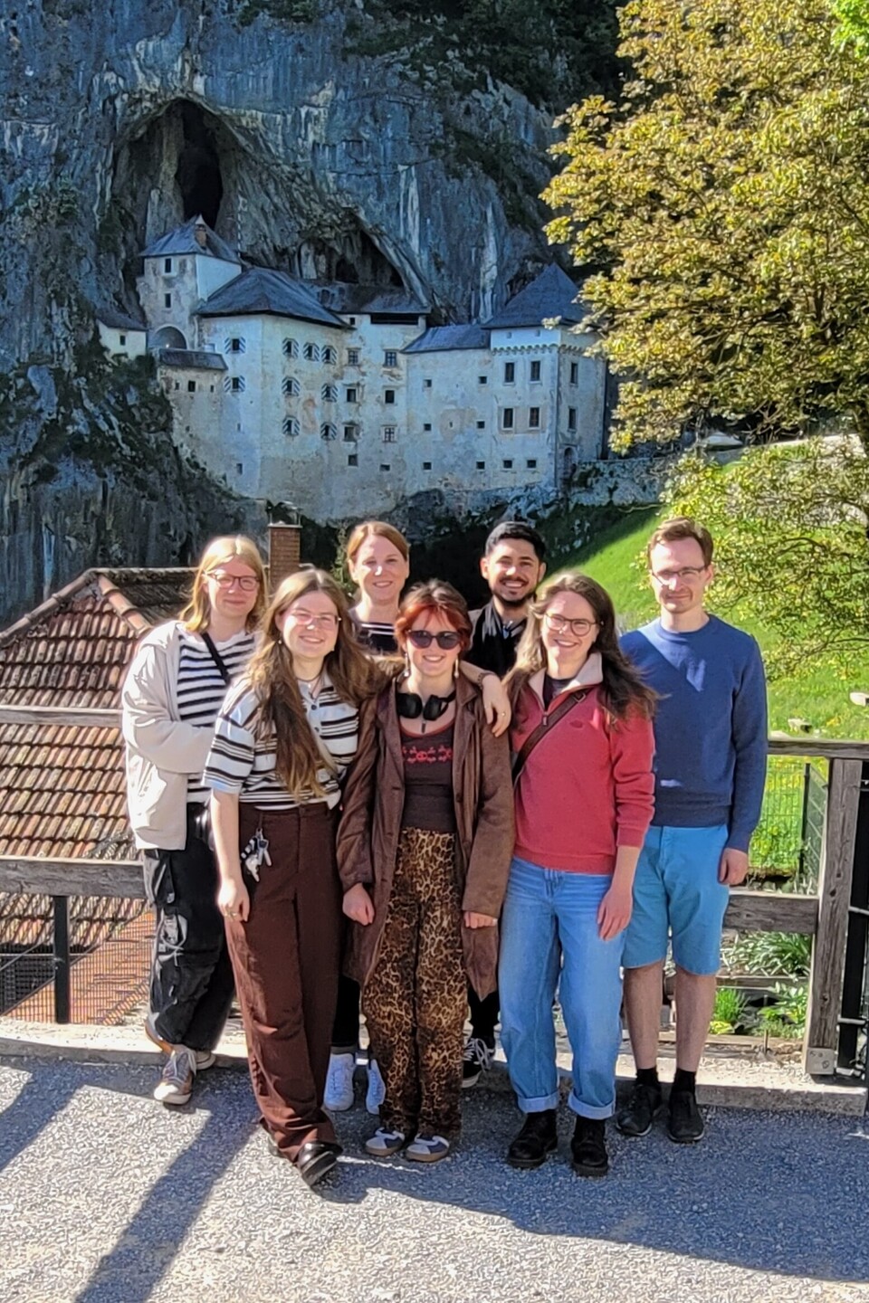 6 Mannheimer Studierende und eine Dozentin stehen vor einer Burg, die vor eine Felsenhöhle gebaut ist, rechts ein Baum und eine grüne Wiese.