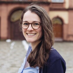 Ein Porträtfoto von der lächelnden Rebekka Hallner. Sie hat braune Haare, eine braune Brille und eine blaue Bluse.