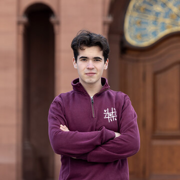 Benedikt trägt einen roten Pullover und schaut seriös mit verschränkten Armen in die Kamera.