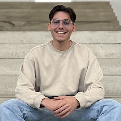 Fabian Herdtle is sitting on a staircase, laughing. He has short hair, is wearing a beige sweater, and large glasses.