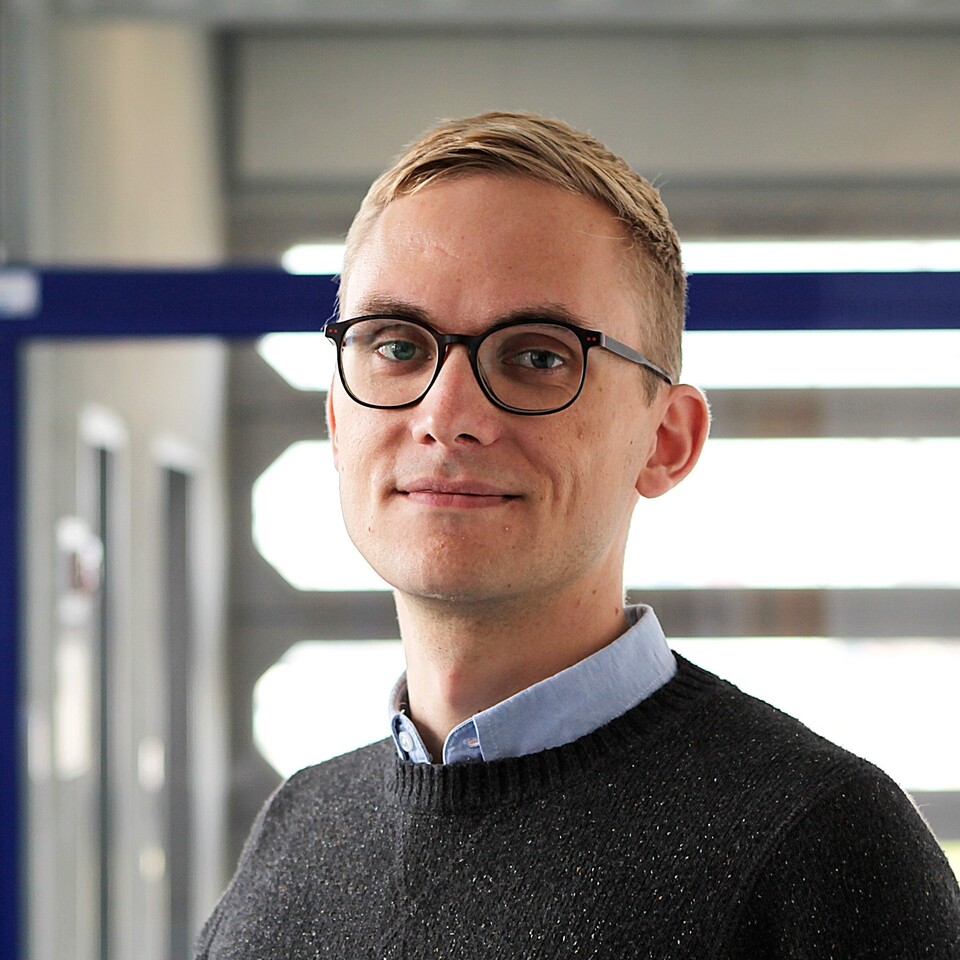 Ruben Bach steht in einem hellen Flur vor Glastüren. Er hat kurze blonde Haare, trägt eine Brille und einen dunklen Pullover.