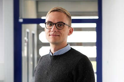 Ruben Bach steht in einem hellen Flur vor Glastüren. Er hat kurze blonde Haare, trägt eine Brille und einen dunklen Pullover.