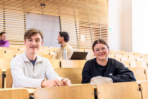 Studierende sitzen in einem Hörsaal. Sie lächeln in die Kamera.