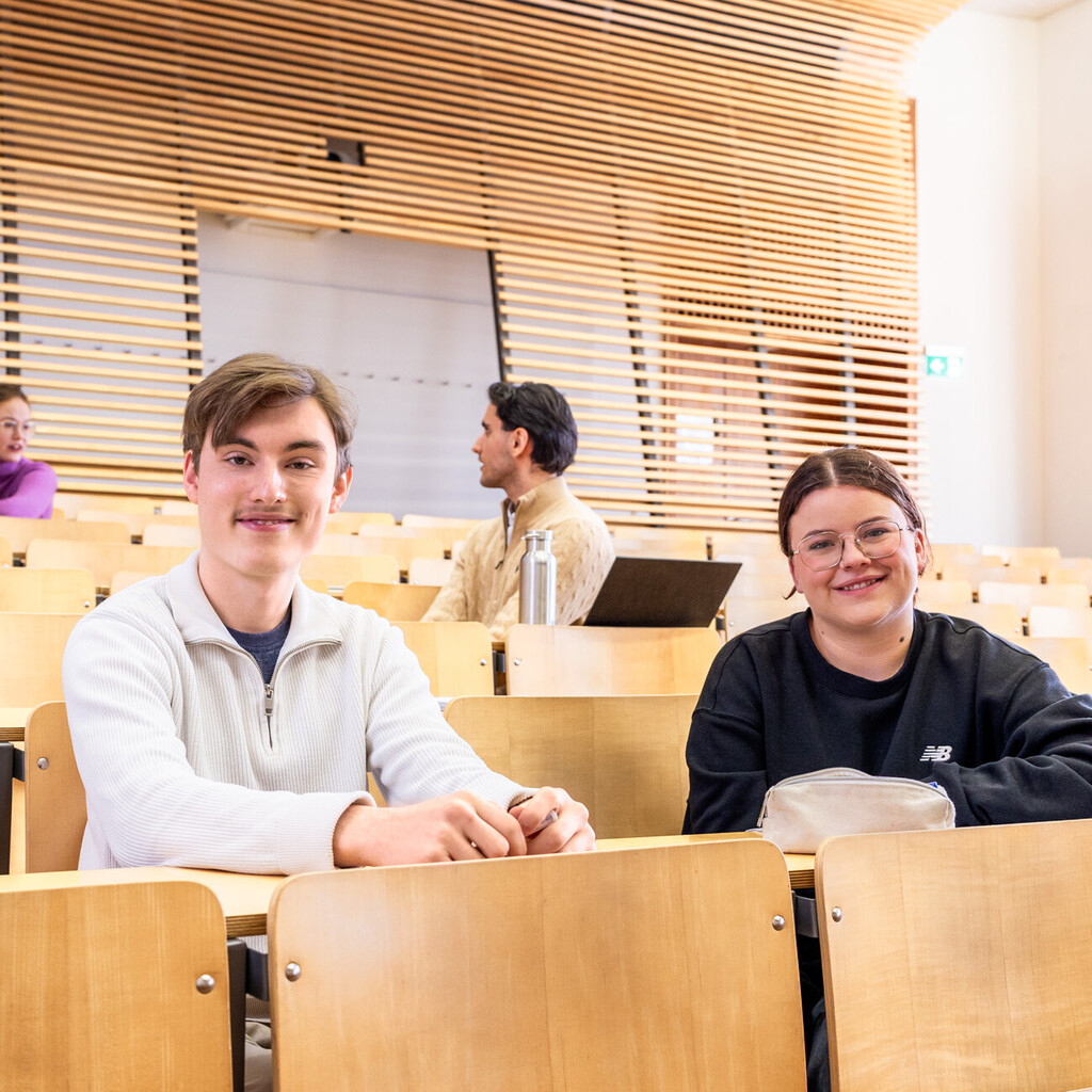 Studierende sitzen in einem Hörsaal. Sie lächeln in die Kamera.