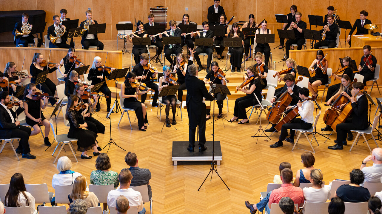 Auftritt des Uniorchesters. Dirigent steht vor den Studierenden, die jeweils ein Instrument spielen. Alle sind schwarz gekleidet.