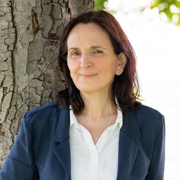 Frau mit schulterlangen braunen Haaren, weißer Bluse, dunkelblauen Blazer und dunkelblauer Hose steht auf der Mensawiese unter einem Baum und lächelt.