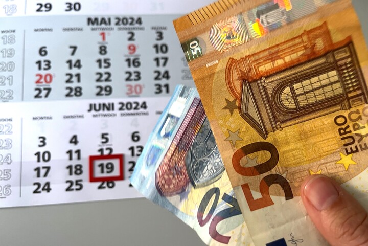 Im Hintergrund liegt ein Kalender, in dem ein Tag rot eingekringelt ist. Im Vordergrund ist eine Hand, die einen 50  Euro Schein und einen 20 Euro Schein hält