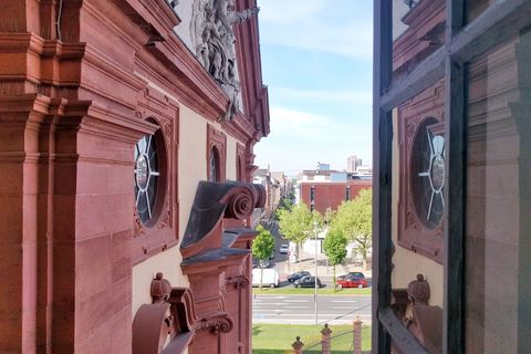 Verzierungen an der Wand des sandsteinfarbenen Mannheimer Schlosses. Ein Fenster ist geöffnet und zeigt eine vielbefahrene, mehrspurige Straße und ein paar Bäume.