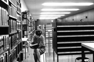 Schwarz-weiß Aufnahme des damaligen Katalogsaals. Eine weiblich gelesene Studentin sitzt vor einem Bücherregal und liest ein Buch.