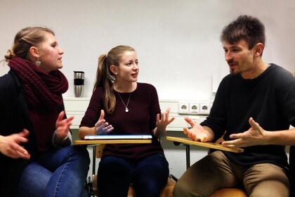 Zwei Frauen und ein Mann sitzen auf Stühlen in einem Halbkreis. Sie diskutieren miteinander und gestikulieren dabei mit ihren Händen.