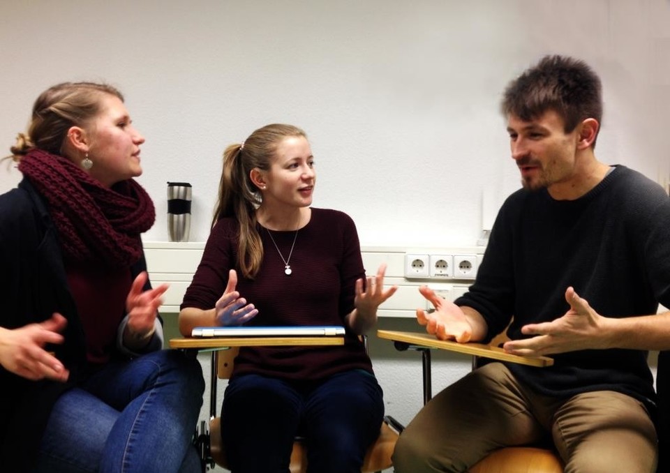 Zwei Frauen und ein Mann sitzen auf Stühlen in einem Halbkreis. Sie diskutieren miteinander und gestikulieren dabei mit ihren Händen.