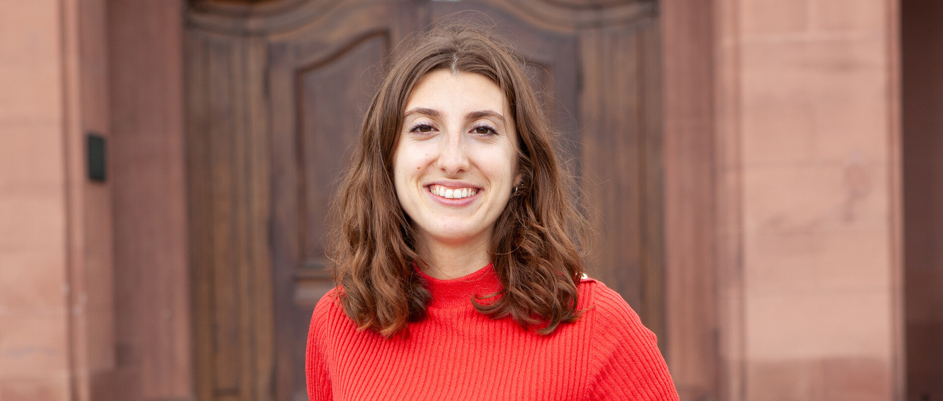 Portrait von Lea Beringuer. Sie trägt ein rotes T-Shirt und steht lächelnd vor dem Schloss.