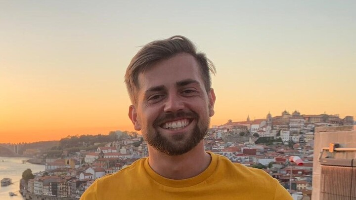 Samuel Mäurer. He is wearing a tangerine shirt and standing in front of a coastal landscape.