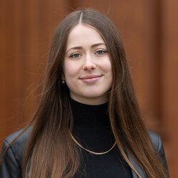 Portrait of Lenja Seubert. She is wearing a black turtleneck sweater and a black leather jacket. Her long brown hair is loose over her shoulders. She is smiling at the camera.