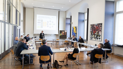 Vierzehn Personen in schicker Kleidung sitzen in einem Konferenzraum und sprechen miteinander.